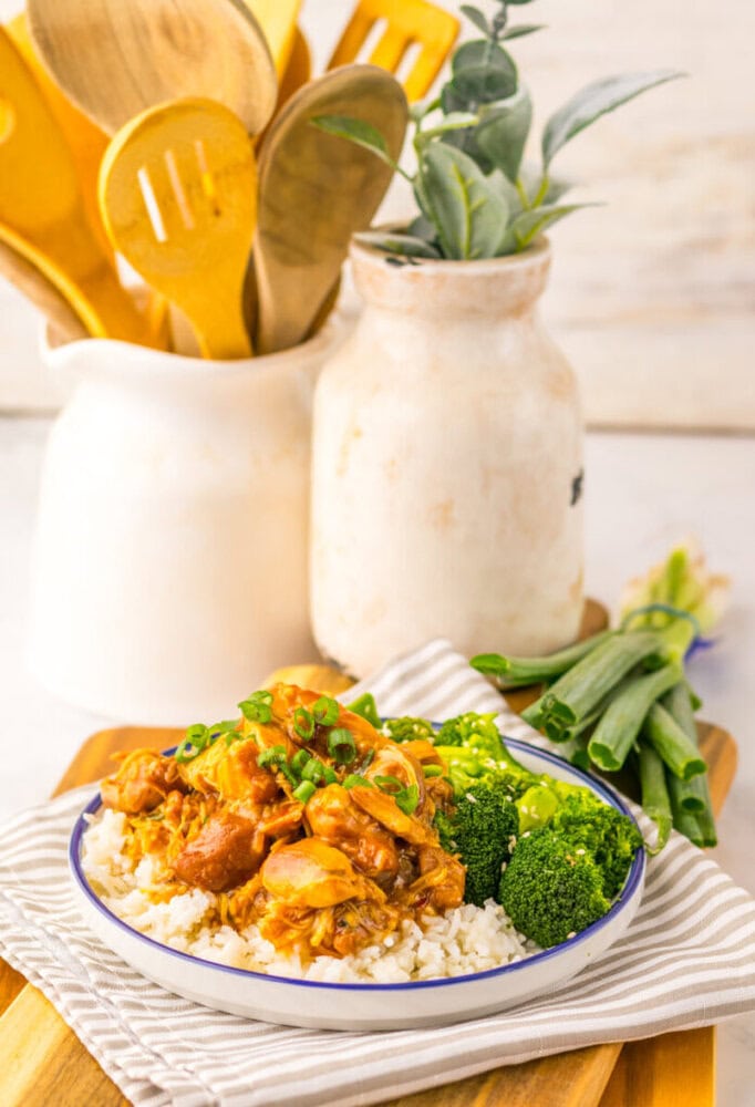 A plate of rice topped with saucy chicken pieces, chopped green onions, and steamed broccoli, placed on a striped cloth with green onions and kitchen utensils in the background.