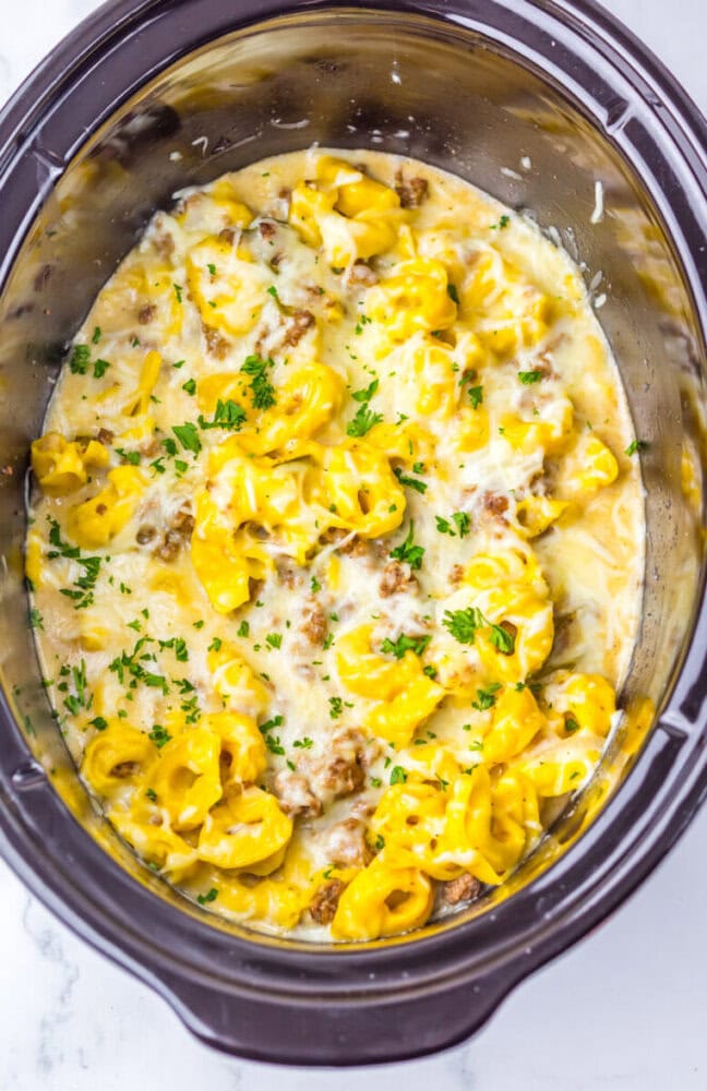 Creamy tortellini with sausage and melted cheese garnished with chopped parsley inside a slow cooker.