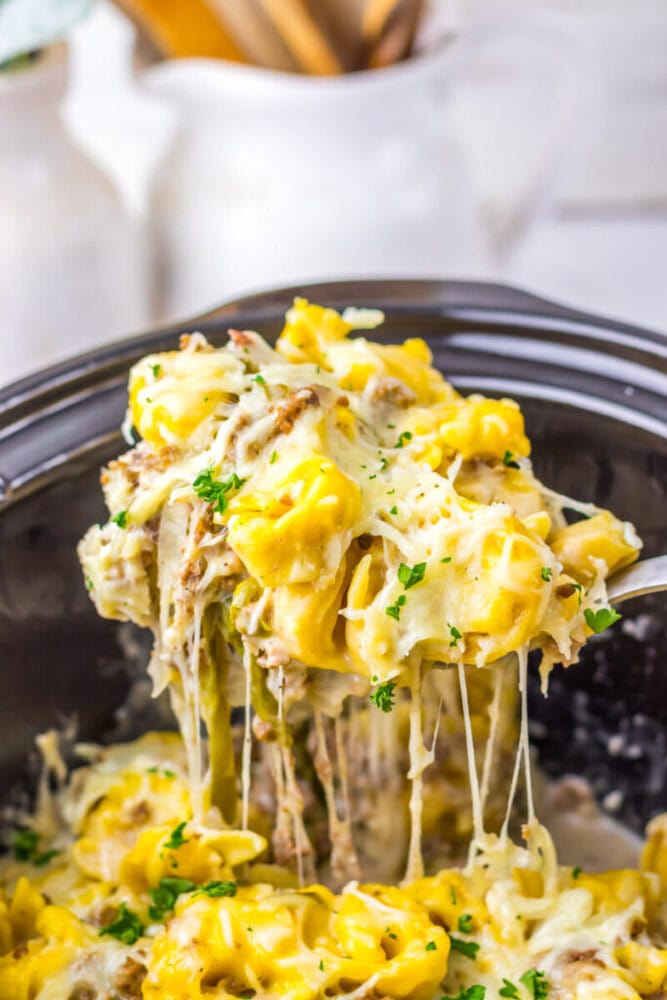 A spoon lifts cheesy tortellini mixed with herbs and shredded meat from a slow cooker, with melted cheese stretching above the pot.