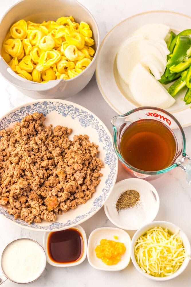 Ingredients for a recipe arranged on a table, including tortellini, sliced onion and green pepper, cooked ground meat, broth, shredded cheese, cream, sauces, and seasonings in bowls and a measuring cup.