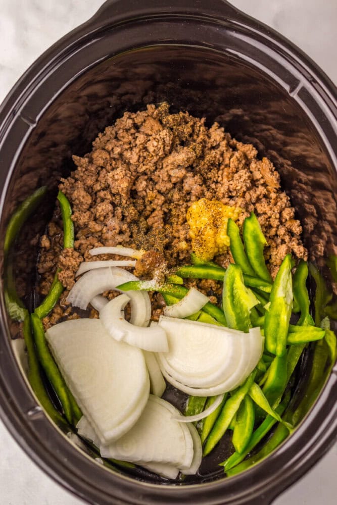 Ground beef, sliced onions, and green bell peppers with seasonings are placed in a slow cooker, ready to be cooked.