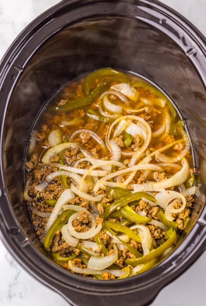 Ground meat, green bell pepper strips, and sliced onions cooked in broth inside a black slow cooker.