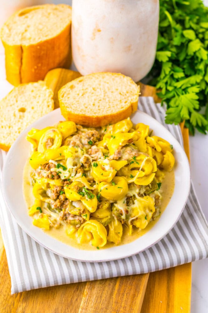 A plate of creamy tortellini with ground meat, garnished with parsley, served with slices of bread on a striped cloth, with fresh parsley in the background.
