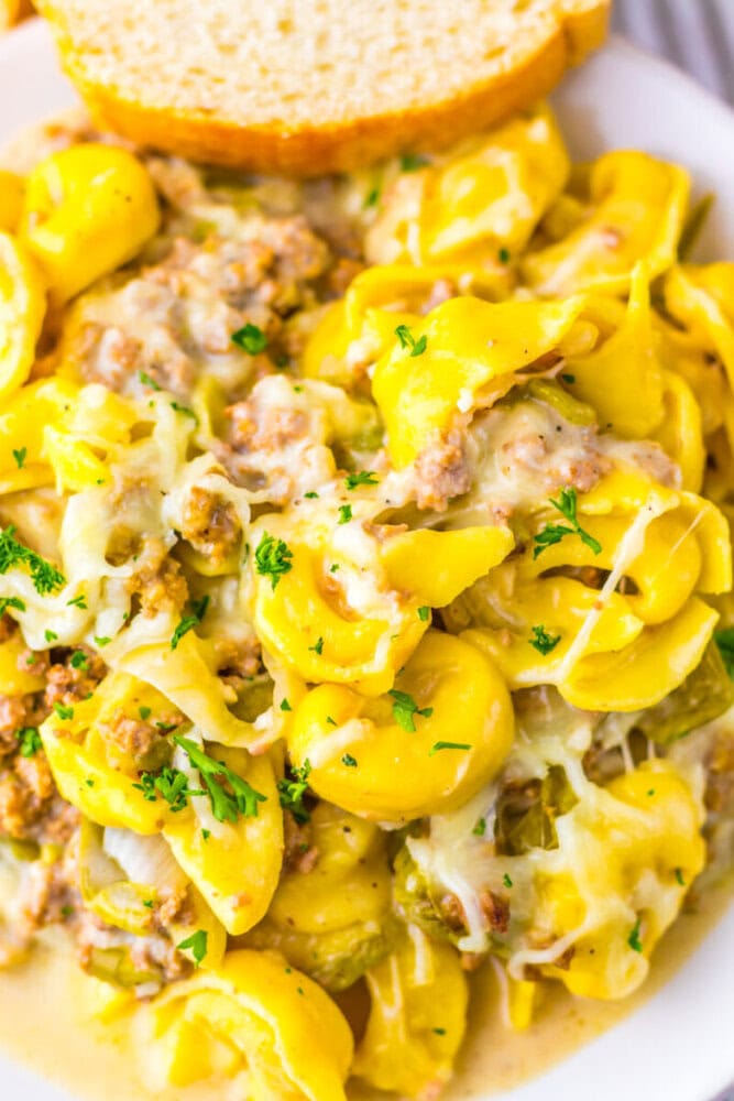 A close-up of cheesy tortellini pasta with ground meat and herbs, served on a plate with a slice of bread in the background.