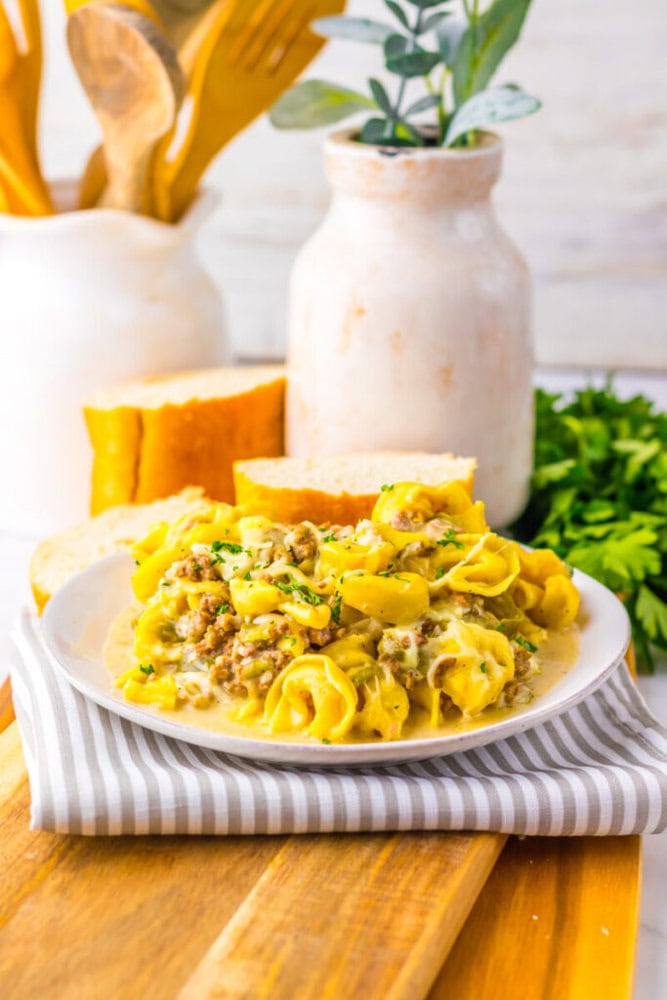 A plate of creamy tortellini with ground sausage is served with slices of bread, set on a striped napkin with a wooden board, parsley, and kitchen utensils in the background.