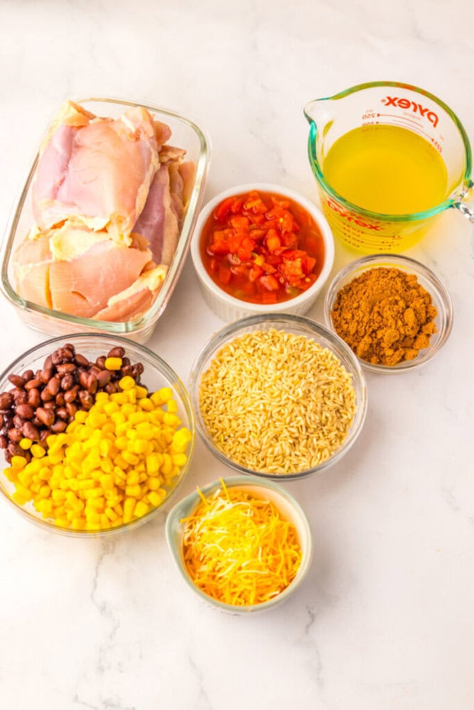 A display of raw chicken thighs, chicken broth, diced tomatoes, taco seasoning, shredded cheese, corn, black beans, and uncooked rice on a white surface.