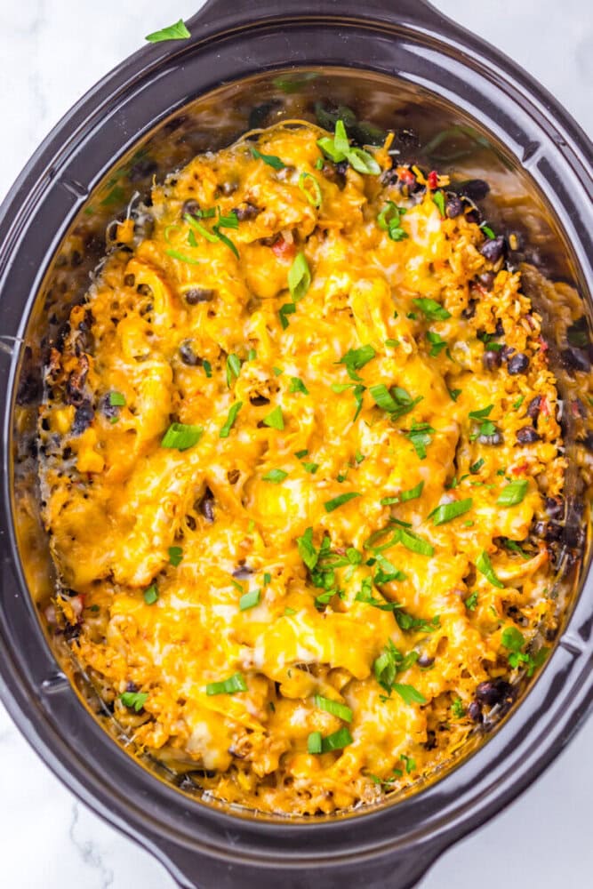 A slow cooker filled with a baked casserole topped with melted cheddar cheese and garnished with chopped green herbs.