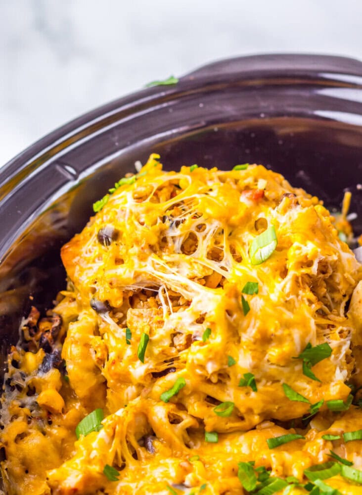 Close-up of a cheesy casserole with shredded chicken and vegetables topped with melted cheese and fresh chopped herbs in a slow cooker.