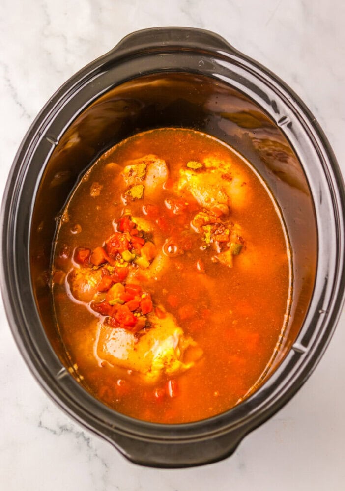 A slow cooker filled with chicken pieces, diced tomatoes, and broth, positioned on a marble countertop.