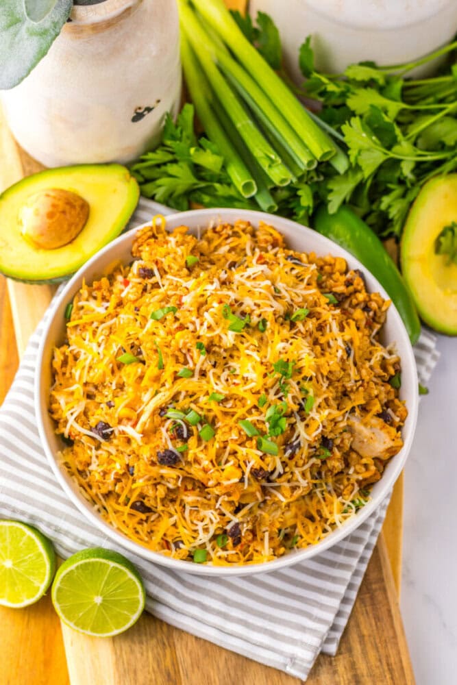 A bowl of cheesy rice with black beans, garnished with green onions and cilantro, surrounded by avocado halves, limes, celery, and fresh herbs on a wooden board.