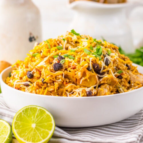 A white bowl filled with Mexican rice, black beans, shredded cheese, and chicken, garnished with cilantro, with lime halves and avocado in the background.