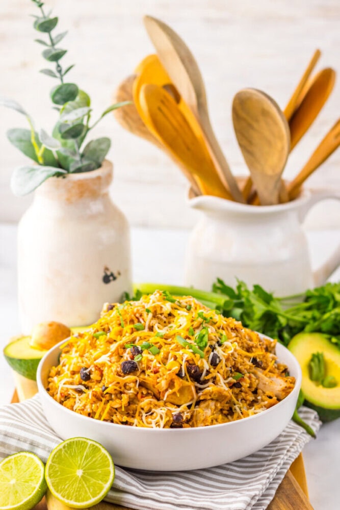 A bowl of rice mixed with vegetables and cheese, garnished with green onions, sits on a striped cloth next to sliced avocado, lime halves, and kitchen utensils in the background.