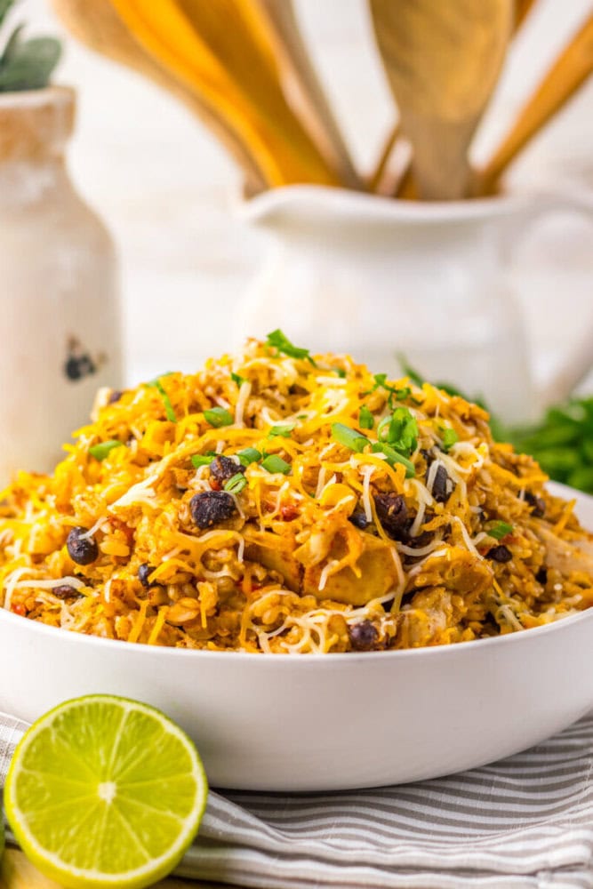 A bowl of shredded chicken and rice dish topped with cheese and herbs, with black beans visible and a halved lime on the side.