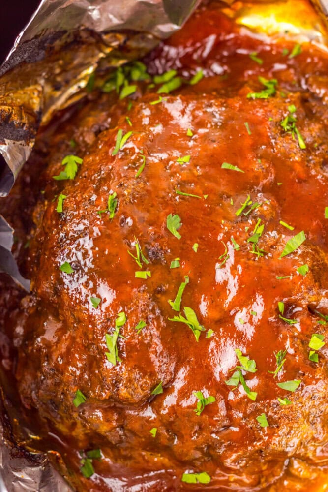 A close-up of a glazed meatloaf topped with chopped parsley, wrapped in foil.