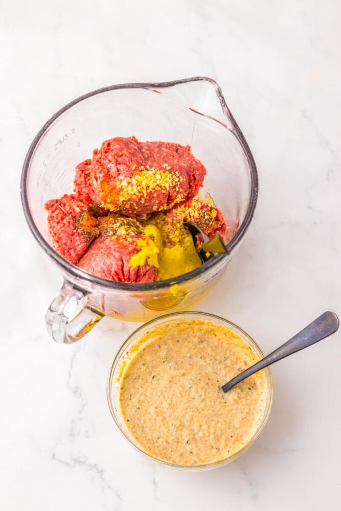 A glass measuring cup with raw ground beef, seasonings, and egg, next to a small bowl of breadcrumb mixture with a spoon, on a white surface.