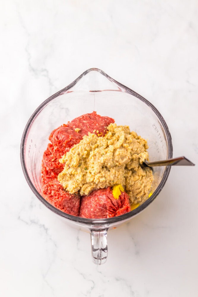 Ground meat, a yellow ingredient, and a beige mixture are in a glass mixing bowl with a spoon, on a white marble surface.
