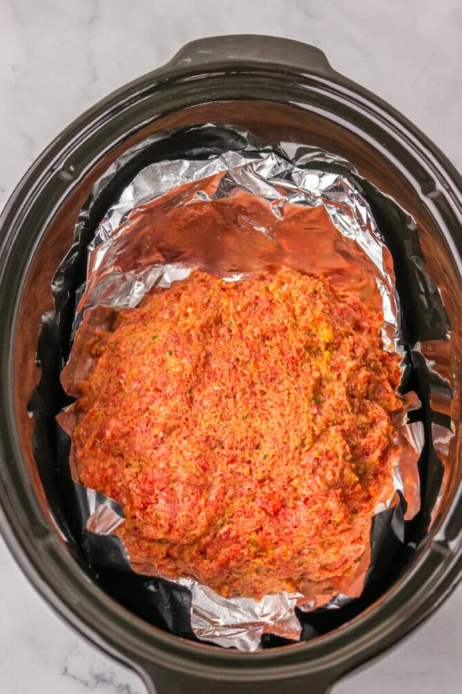 Uncooked meatloaf mixture shaped and placed on a sheet of aluminum foil inside a slow cooker on a marble countertop.