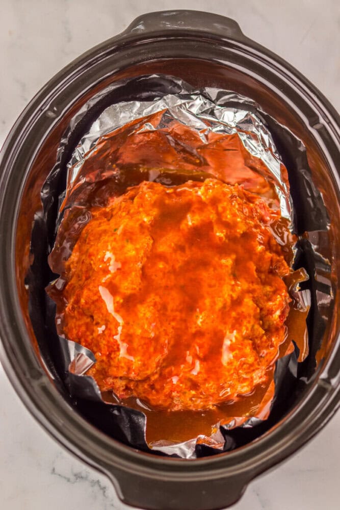 A raw, seasoned meatloaf covered in sauce sits on aluminum foil inside a slow cooker on a marble countertop.