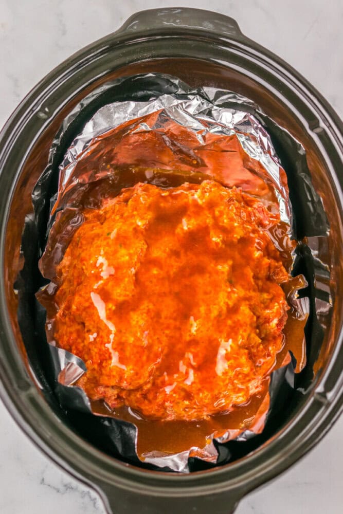 A slow cooker lined with aluminum foil contains a mound of raw ground meat covered in a red sauce.
