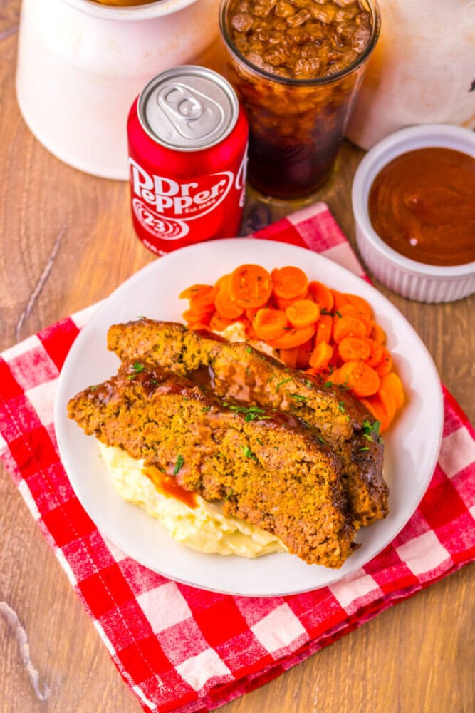 A plate with two slices of meatloaf on mashed potatoes, sliced carrots, a can and glass of Dr Pepper, and a small dish of sauce on a red checkered napkin.