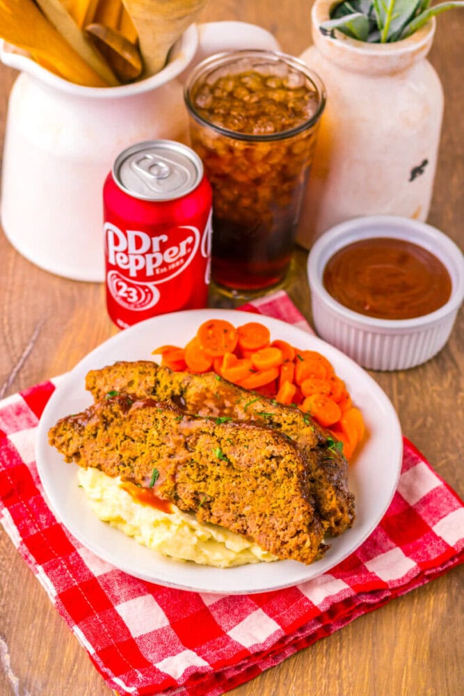 A plate of meatloaf on mashed potatoes with sliced carrots, next to a ramekin of sauce, a can and glass of Dr Pepper, on a red checkered cloth.