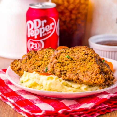 A plate with two slices of meatloaf on mashed potatoes sits on a red checkered cloth, with a can of Dr Pepper and a glass of iced drink in the background.