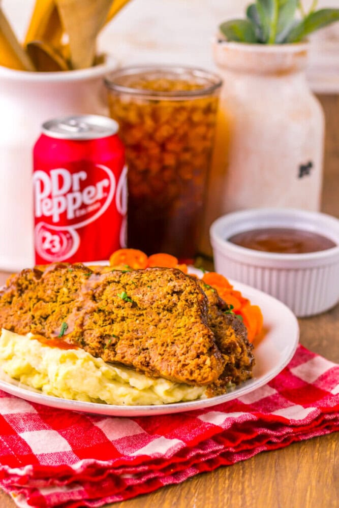 A plate with two slices of meatloaf on mashed potatoes, sliced carrots, a bowl of sauce, a can of Dr Pepper, and a glass of iced drink on a red checkered cloth.