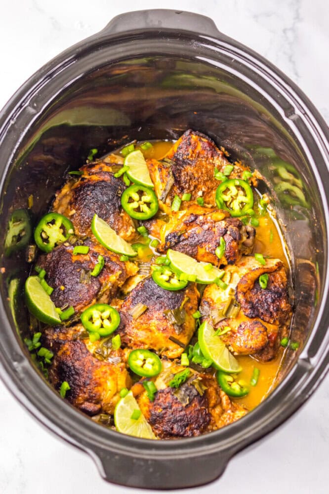 Slow cooker filled with cooked chicken thighs in sauce, garnished with sliced jalapeños, lime wedges, and chopped herbs.