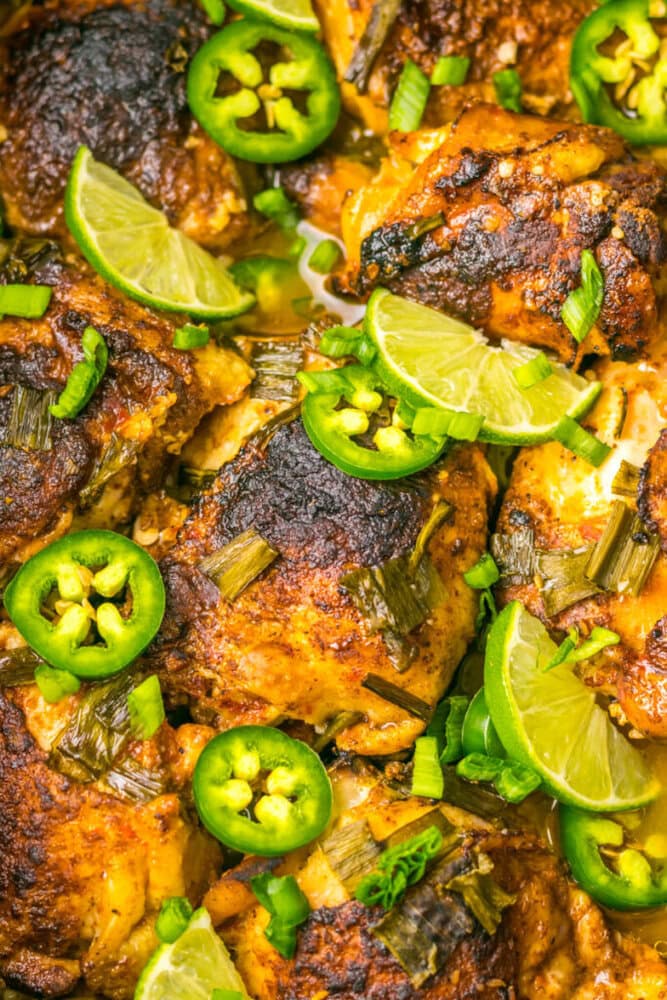 Close-up of seasoned, roasted chicken thighs garnished with lime wedges, sliced jalapeños, and green onions.