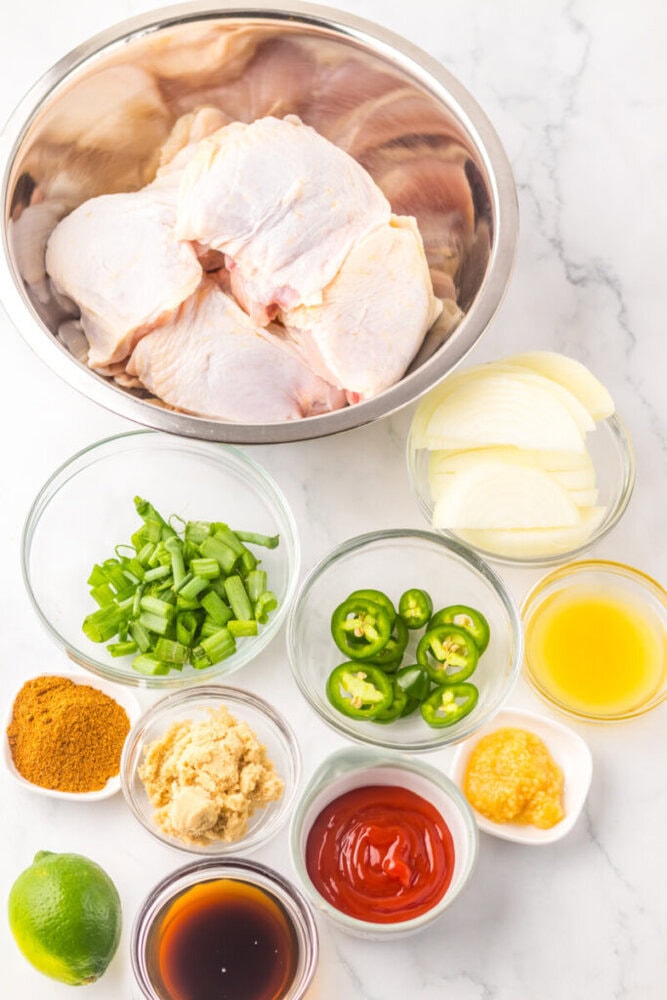 Raw chicken pieces in a metal bowl, surrounded by small bowls of sliced onions, green onions, jalapeños, spices, ginger, lime, ketchup, soy sauce, and orange juice on a marble surface.