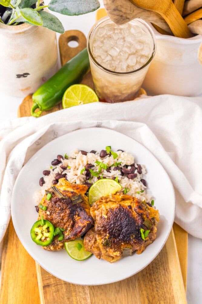 A plate of rice and black beans topped with stewed chicken, garnished with lime and jalapeño slices, next to a glass of iced drink and a green pepper.