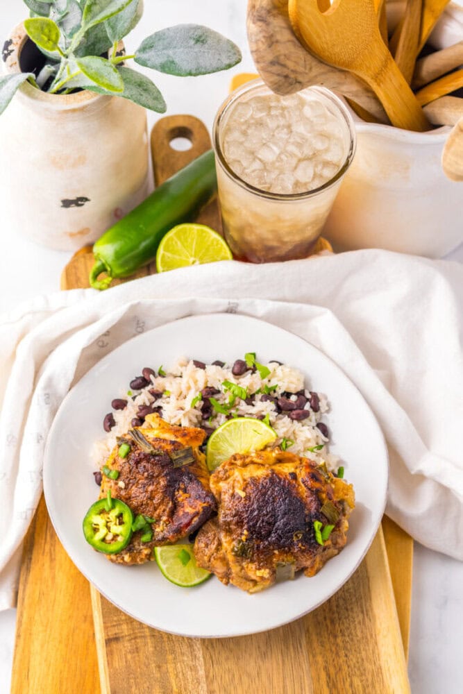 A plate of rice and beans topped with two pieces of seasoned chicken, garnished with lime and sliced jalapeño, with a glass of iced drink and a green pepper in the background.