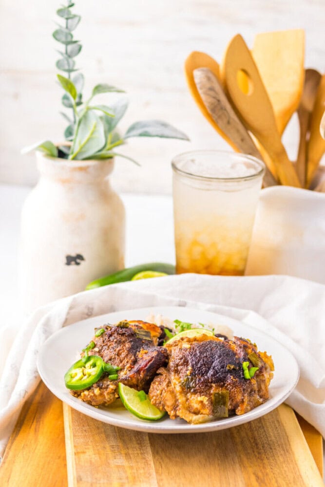 A plate with two pieces of cooked chicken garnished with sliced jalapeños and green onions, set on a wooden board with a cold drink, vase, and utensils in the background.