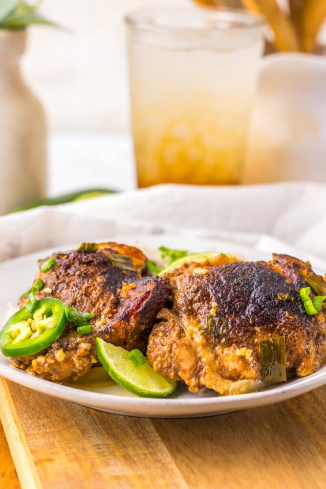 Two pieces of seasoned, cooked chicken topped with sliced green jalapeños and garnished with green onions on a white plate. A drink with ice is in the background.