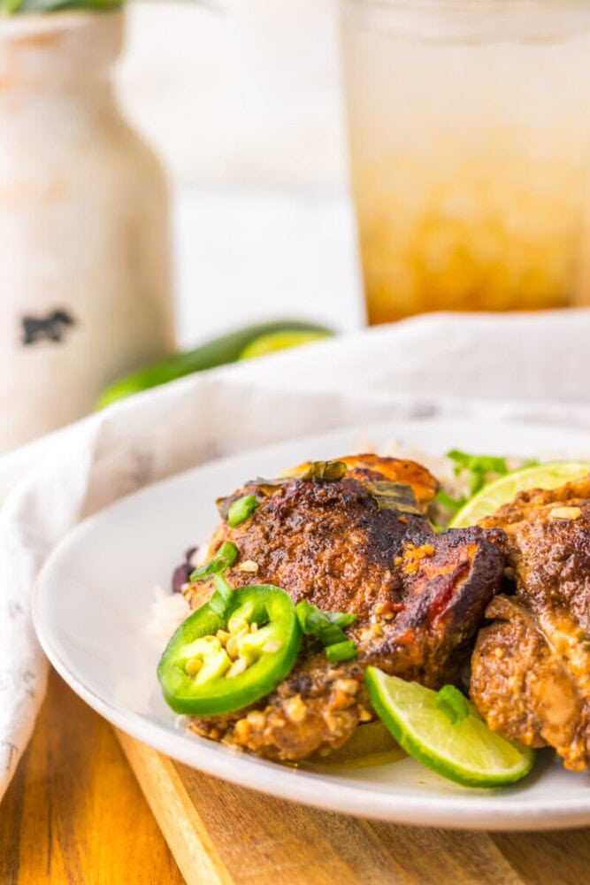 A plate of cooked seasoned chicken topped with sliced jalapeños and green onions, garnished with lime wedges, sits on a table.