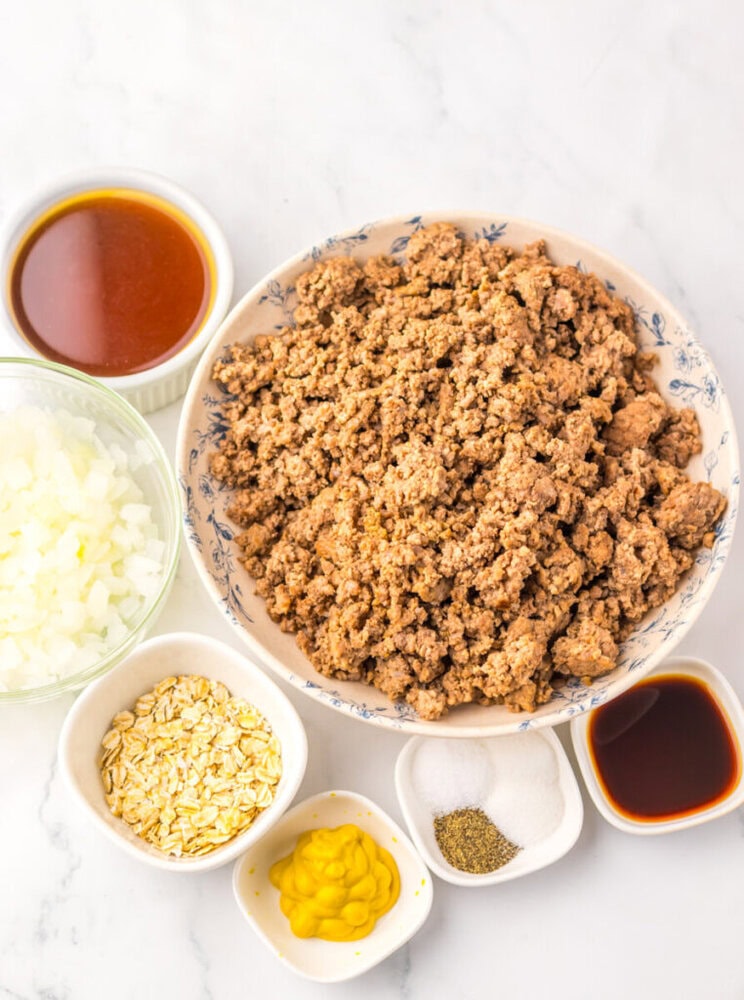 A bowl of cooked ground beef surrounded by bowls of chopped onions, oats, barbecue sauce, mustard, salt, pepper, and Worcestershire sauce on a white surface.