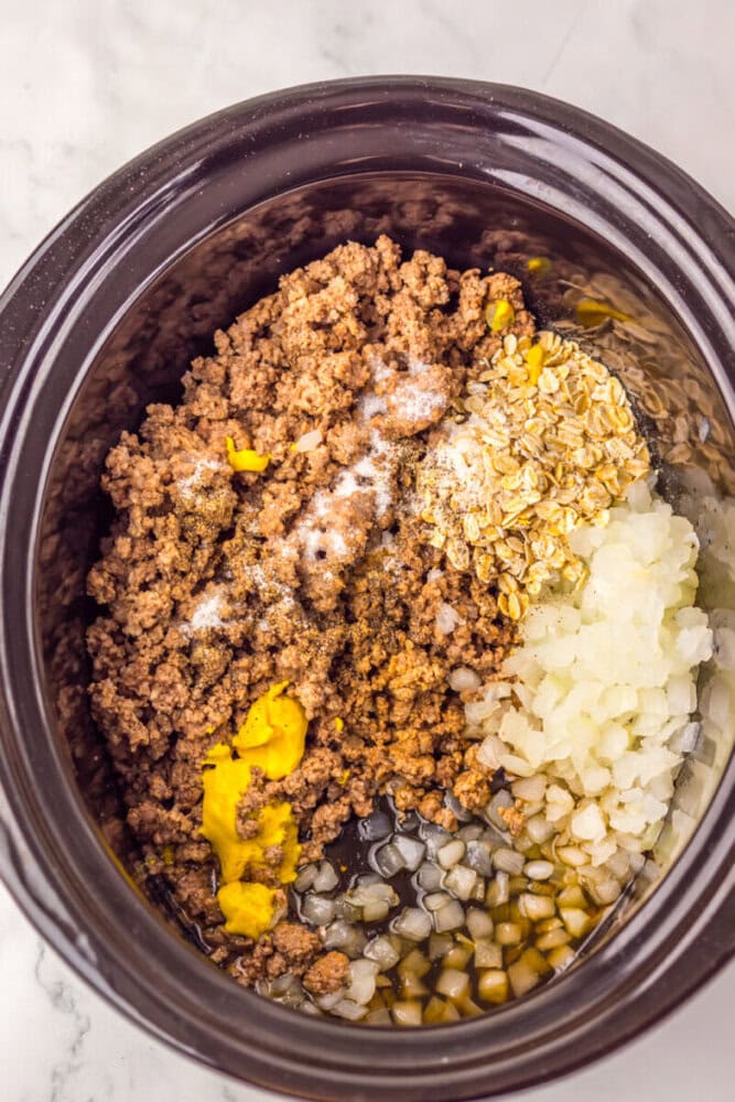 Ground beef, chopped onions, oats, mustard, seasonings, and liquid ingredients in a slow cooker, ready to be mixed.