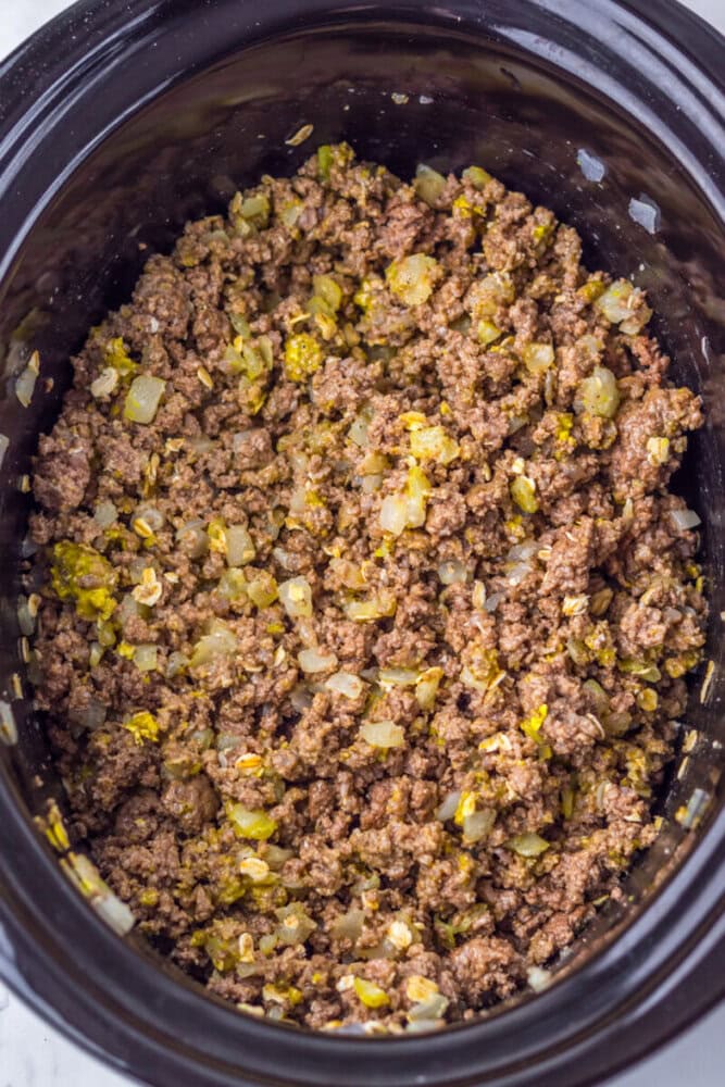 Cooked ground beef mixed with chopped onions and seasonings in a black slow cooker.