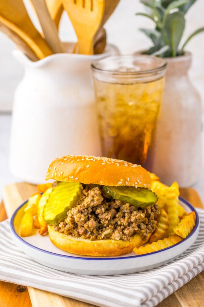 A sloppy joe sandwich with pickles on a sesame bun, served with crinkle-cut fries and a drink, sits on a plate on a wooden board.