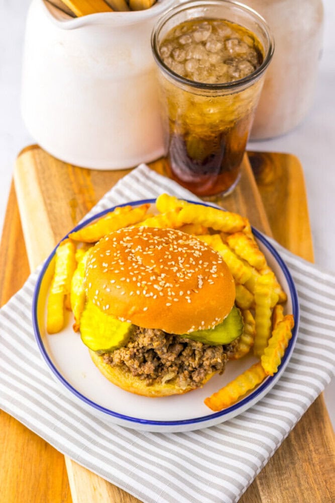 A plate with a sloppy joe sandwich topped with pickles and crinkle-cut fries, next to a glass of soda on a wooden board.