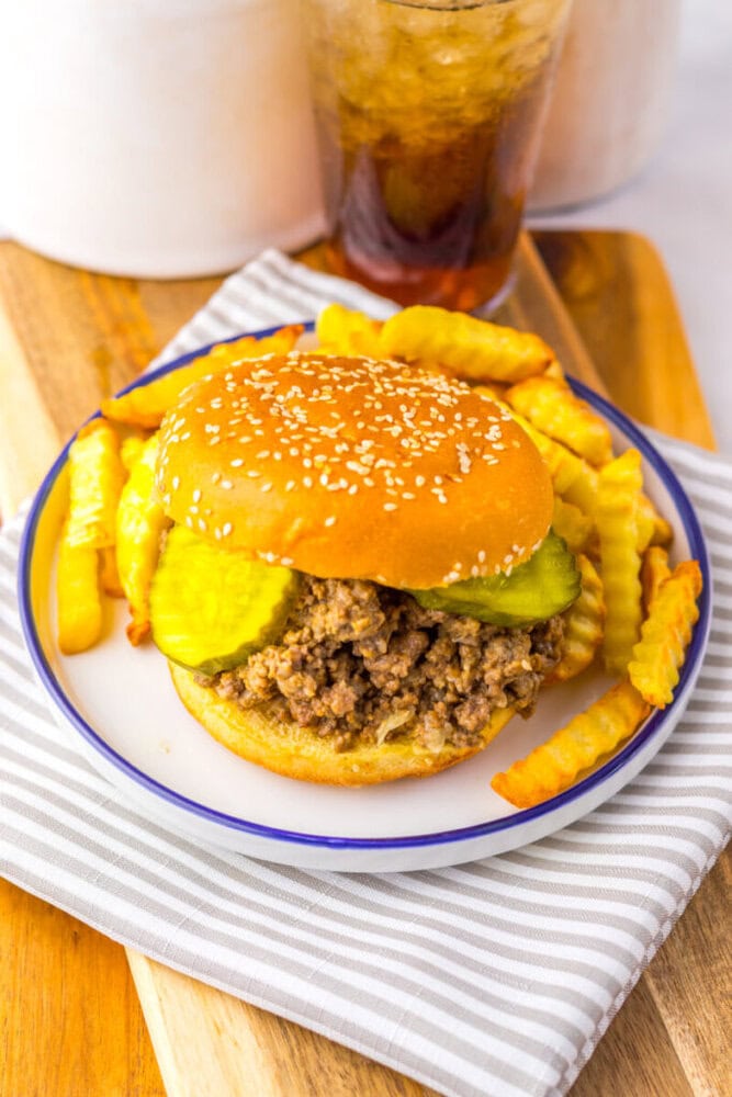 A sloppy joe sandwich with pickles and crinkle-cut fries on a plate, next to a glass of soda on a wooden surface.