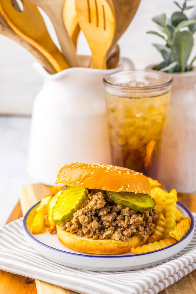A sloppy joe sandwich with pickles on a sesame bun, served with crinkle-cut fries and a drink on a plate, with wooden utensils and a potted plant in the background.