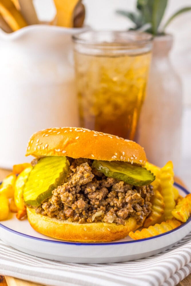A sloppy joe sandwich with pickle slices on a sesame bun, served with crinkle-cut fries and a glass of iced drink in the background.