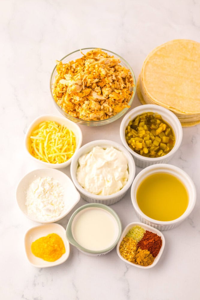 Assorted ingredients in small bowls on a white surface, including shredded chicken, shredded cheese, sour cream, green chilies, oil, flour, milk, spices, and corn tortillas.
