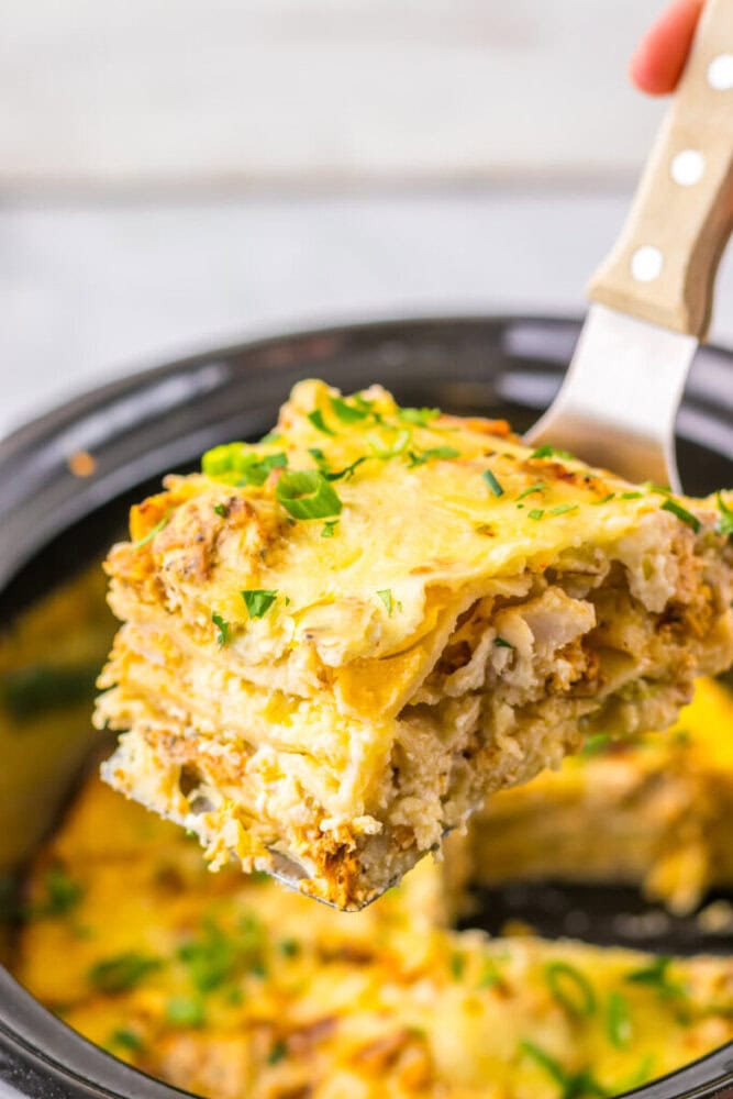 A spatula lifts a piece of cheesy, layered lasagna topped with chopped herbs from a slow cooker.