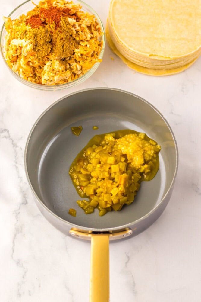 A saucepan with chopped green chilies, a bowl of shredded chicken with spices, and a stack of corn tortillas on a marble surface.