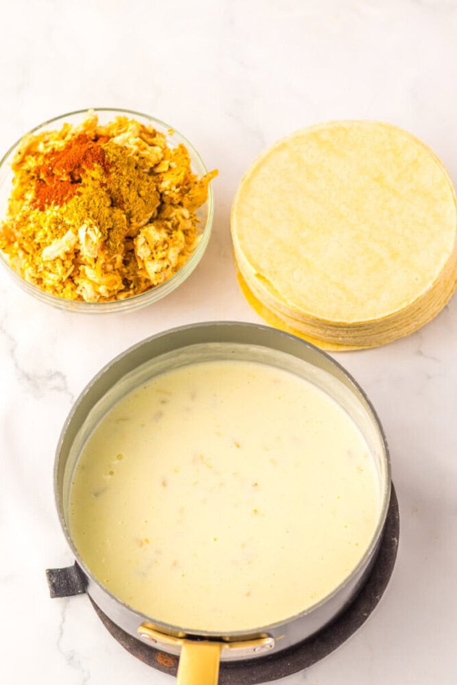 A pot of creamy sauce, a bowl of shredded chicken with spices, and a stack of corn tortillas on a white surface.