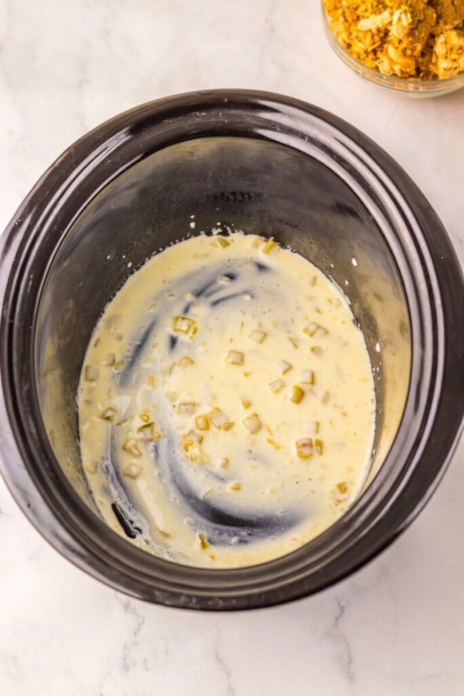 A slow cooker containing a creamy mixture with chopped pickles on a light-colored countertop.