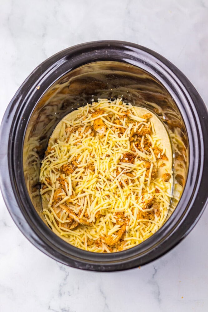 A slow cooker with shredded cheese and ground meat layered inside, ready for cooking, on a white marble surface.