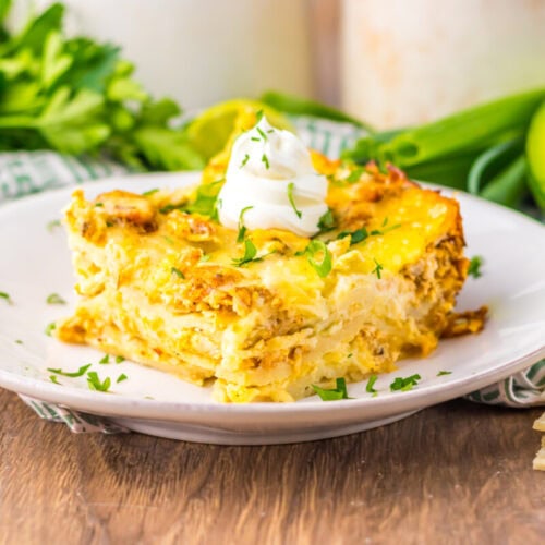 A slice of egg casserole topped with sour cream and chopped herbs on a white plate, with avocado, tortillas, and fresh greens in the background.